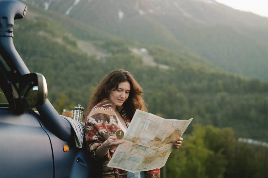 Woman wrapped in a patterned blanket reading a map and holding a compass beside a car in the mountains.
