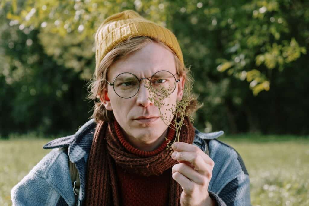 A person wearing glasses and a yellow beanie is closely examining a wild plant outdoors.