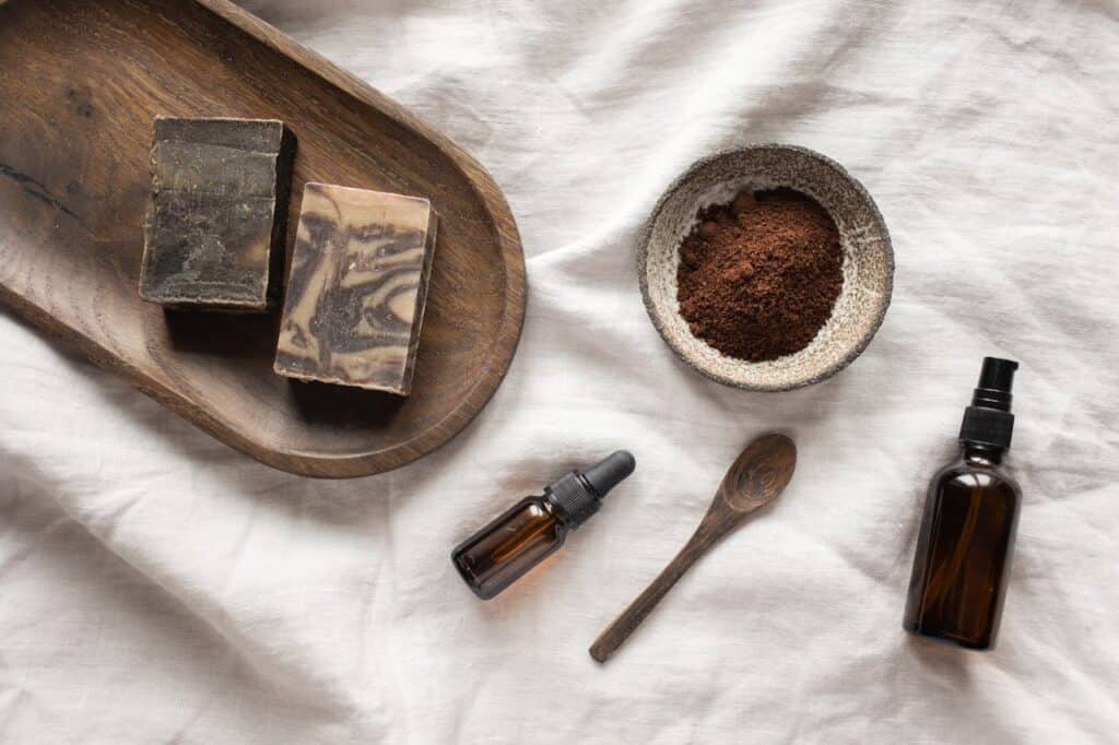 Natural soaps, a bowl of ground coffee, glass bottles, and a wooden spoon on a white cloth